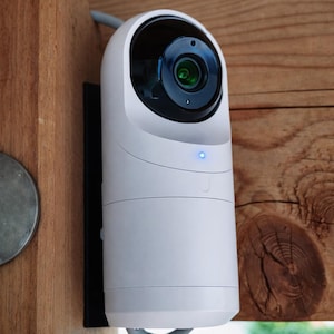 May include: A white security camera with a black lens and a blue indicator light is mounted on a wooden post. The camera is connected to a gray cable and is designed for outdoor use.