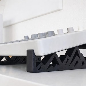 以下が含まれることがあります： グレーのノブが付いた白い電子機器が、黒い幾何学的なスタンドの上に置かれています。スタンドは山のようなデザインです。デバイスは上向きに傾斜しており、キーボードまたはコントロールパネルを示唆しています。