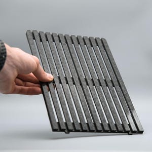 May include: A dark gray, rectangular grill grate with evenly spaced, vertical slats. The grate is held by a hand with a black watch. The background is a solid, light gray.