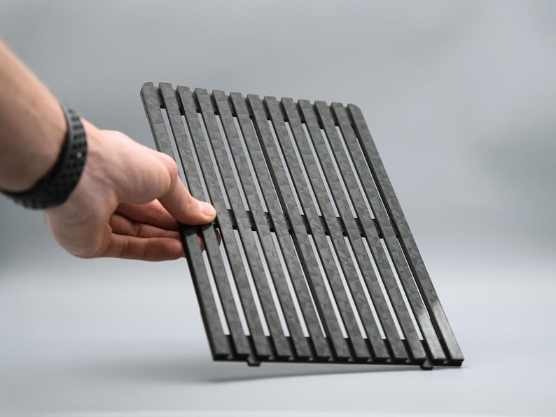 May include: A dark gray, rectangular grill grate with evenly spaced, vertical slats. The grate is held by a hand with a black watch. The background is a solid, light gray.