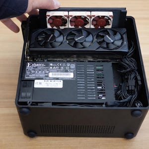 Peut inclure: Boîtier d'ordinateur noir avec des composants internes visibles. Une main soulève le panneau supérieur pour révéler une carte graphique avec trois ventilateurs noirs et une alimentation. Le boîtier a un extérieur noir et une série d'aérations.