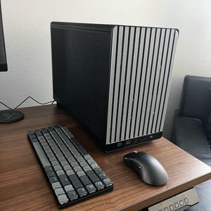 May include: A black and silver computer case with a vertical striped design sits on a wooden desk. A gray keyboard and mouse are positioned in front of the case. The desk has a control panel for height adjustment.