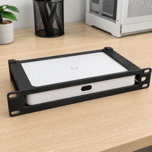 May include: A white Cloud Gateway Fiber network device secured within a black metal rack mount bracket. The device features a small U-shaped logo on the top surface and a black oval port on the front panel, designed for standard server rack installation.