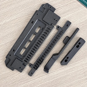 May include: Black, unassembled mechanical device components on a light wood surface. The parts include a large rectangular piece, two slotted bars, and a smaller angled piece. The components appear to be made of a matte black material.
