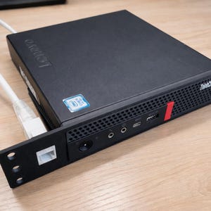 Puede incluir: Un ordenador Lenovo ThinkCentre negro con un cable Ethernet blanco conectado. El ordenador tiene una franja roja y varios puertos en el panel frontal. La pegatina de Intel es visible.