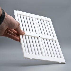 May include: A white plastic vent cover with vertical slats and a rectangular frame. The cover is held by a hand against a gray background. The vent cover is likely used for air conditioning or heating systems.