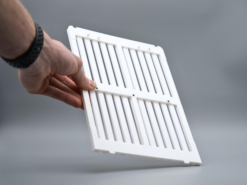 May include: A white plastic vent cover with vertical slats and a rectangular frame. The cover is held by a hand against a gray background. The vent cover is likely used for air conditioning or heating systems.