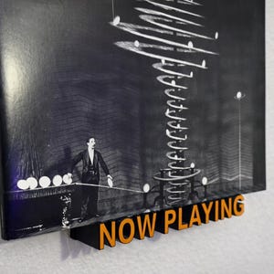 May include: A black and white image of a man in a suit standing in front of a large, spiraling structure. The image is mounted on a wall with a wooden "NOW PLAYING" sign in orange letters.