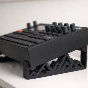 May include: A black electronic music production device with knobs and keys, resting on a black stand. The stand has a geometric mountain-like design. The device is angled upwards, providing an ergonomic setup for the user.