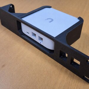 Può includere: Supporto nero stampato in 3D per dispositivi di rete che contiene un dispositivo di rete bianco. Il supporto ha una forma rettangolare con ritagli e fori di montaggio. Il dispositivo di rete ha una parte superiore bianca con un logo curvo e diverse porte.