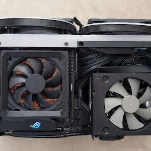 May include: A black computer case interior with multiple fans and components. The case features a large fan with black blades and copper-colored fins, along with other fans and wiring. The ROG logo is visible on a component.