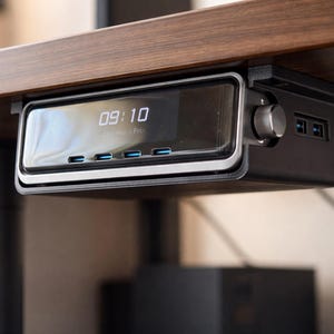 May include: A black and silver desk-mounted charging station with a digital clock displaying 09:10. It has a silver dial, USB ports, and blue-lit charging ports. The station is mounted under a wooden desk.