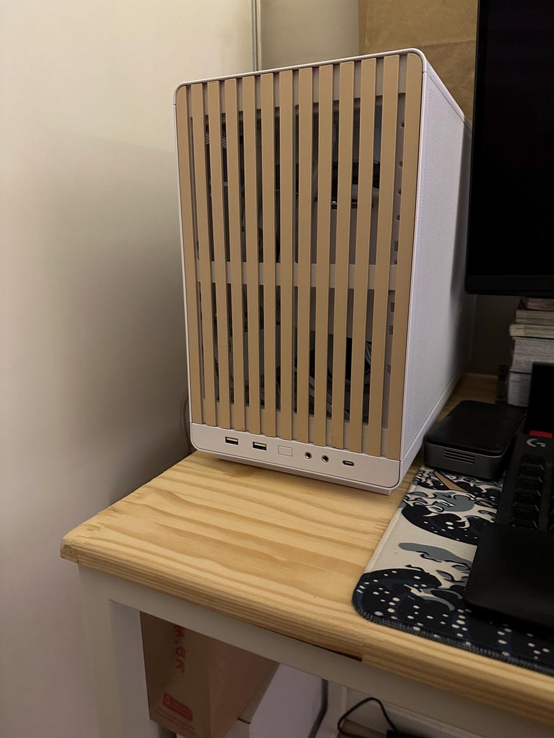 May include: A white computer case with a light brown wooden slat front panel. The case has multiple USB ports and audio jacks on the front. It sits on a light wood table.