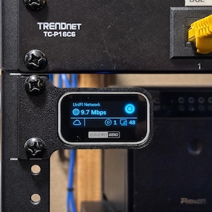 May include: A black UniFi Cloud Key Gen2 network controller mounted on a black metal rack rail. The device features a small digital display showing UniFi Network, 9.7 Mbps, and status icons. Above it, a Trendnet TC-P16C6 patch panel with yellow cables.