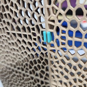 May include: Close-up of a beige, honeycomb-patterned structure with a turquoise clip. The structure has a repeating geometric design, with a blue background visible through the openings. The clip is rectangular and appears to hold two panels together.