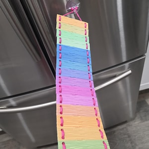 May include: A hanging ladder toy for small animals constructed from rectangular wooden slats in shades of orange, green, blue, and purple. The slats are connected by bright pink nylon cord and suspended from a clear plastic suction cup hook.