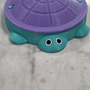 May include: A 3D printed turtle figurine featuring a teal body and a purple shell with a circular grid pattern. The turtle has two white eyes with black pupils and a small smile etched into its face. The shell is secured with small teal fasteners.
