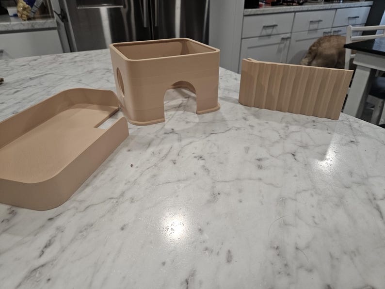 May include: A set of three light beige plastic components for a small animal habitat, including a rectangular tray, a square hideout with an arched entrance and a circular side opening, and a separate ramp piece with a textured, ribbed surface.