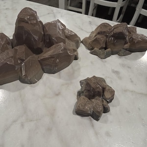 May include: Three brown, angular rock formations of varying sizes, each featuring a faceted, geometric surface texture. These decorative items are sculpted with sharp edges and irregular shapes, resembling clusters of stones or craggy mountain terrain.