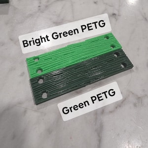 May include: Two rectangular 3D printed plastic components with a wood grain texture and four circular holes each. One component is bright green with a label reading Bright Green PETG, and the other is dark green with a label reading Green PETG.