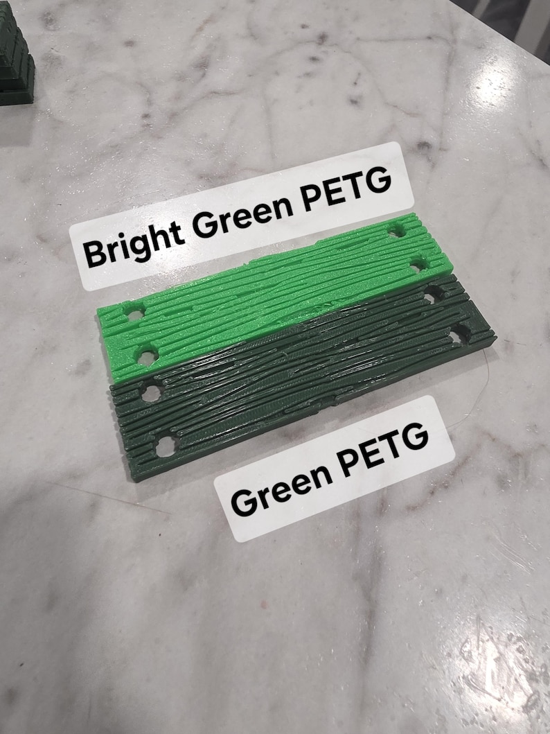 May include: Two rectangular 3D printed plastic components with a wood grain texture and four circular holes each. One component is bright green with a label reading Bright Green PETG, and the other is dark green with a label reading Green PETG.