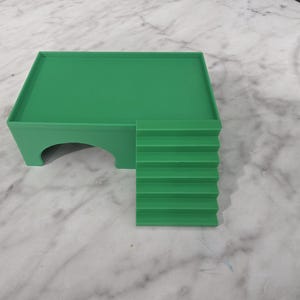 Puede incluir: Una plataforma verde s&oacute;lida con una superficie superior rectangular plana, una &uacute;nica abertura arqueada en el lateral y un conjunto de seis escalones integrados que conducen al nivel de la plataforma. La estructura es de material liso y mate.