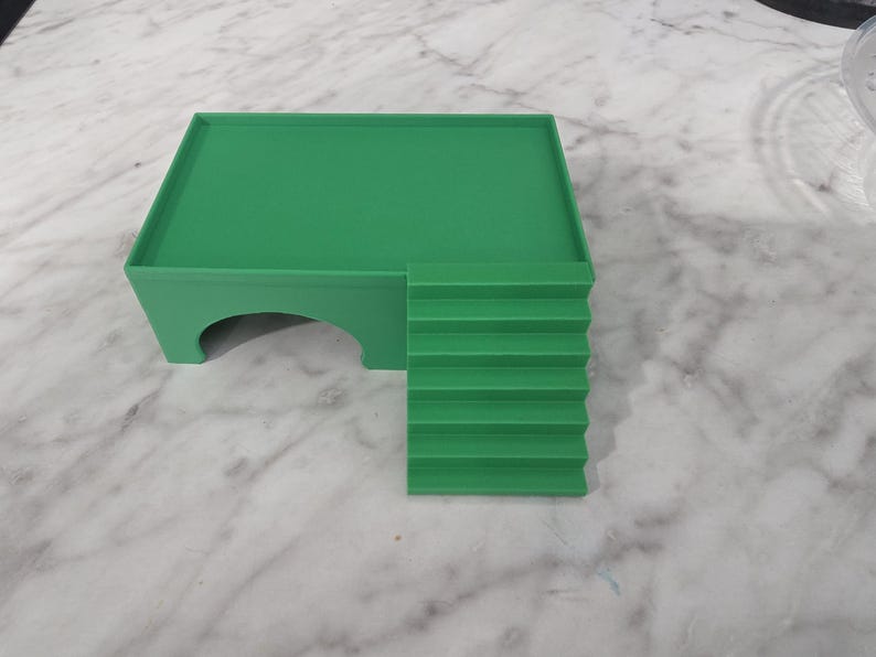 Puede incluir: Una plataforma verde s&oacute;lida con una superficie superior rectangular plana, una &uacute;nica abertura arqueada en el lateral y un conjunto de seis escalones integrados que conducen al nivel de la plataforma. La estructura es de material liso y mate.
