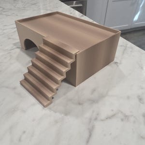 Puede incluir: Una plataforma rectangular de color beige con una superficie superior plana y un borde elevado. La estructura presenta una abertura arqueada en un lado y una escalera de seis pelda&ntilde;os que conduce al nivel de la plataforma, en material mate.
