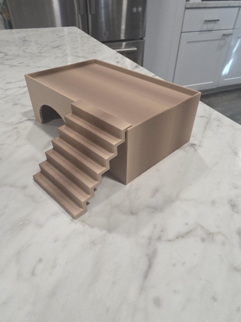 Puede incluir: Una plataforma rectangular de color beige con una superficie superior plana y un borde elevado. La estructura presenta una abertura arqueada en un lado y una escalera de seis pelda&ntilde;os que conduce al nivel de la plataforma, en material mate.