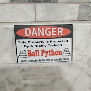 May include: A black and white sign with a red banner that reads "DANGER". The sign says "This property is protected by a highly trained Ball Python. Not responsible for injury or possible death."
