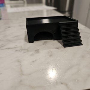 Puede incluir: Un refugio para animales negro, rectangular e impreso en 3D, que cuenta con una plataforma superior plana, una entrada en arco semicircular en la parte frontal y un conjunto de escaleras integradas que conducen a la plataforma en el lado derecho.