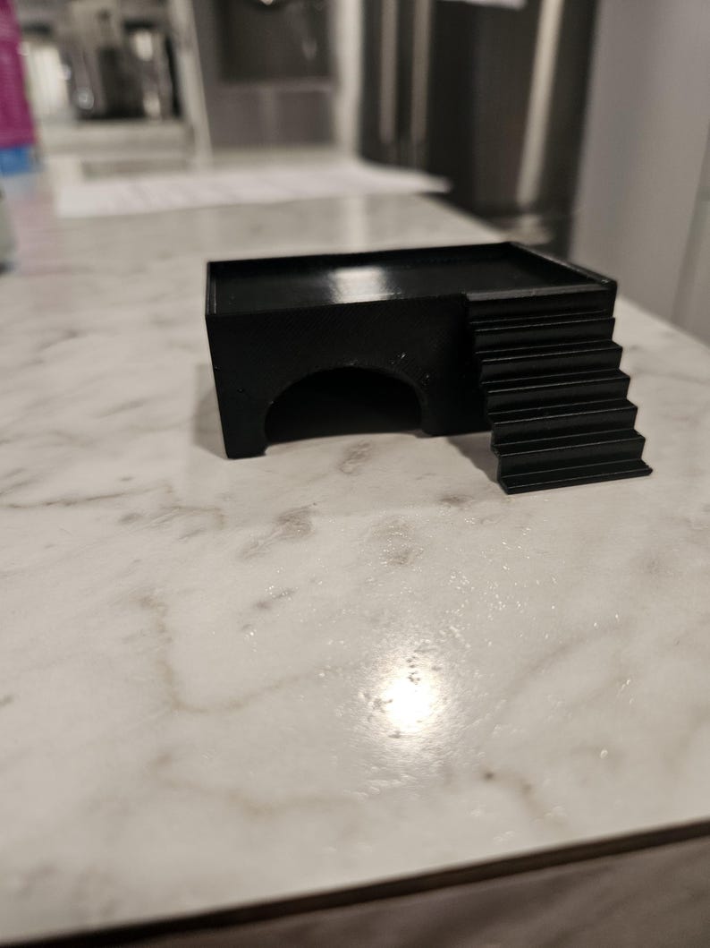 Puede incluir: Un refugio para animales negro, rectangular e impreso en 3D, que cuenta con una plataforma superior plana, una entrada en arco semicircular en la parte frontal y un conjunto de escaleras integradas que conducen a la plataforma en el lado derecho.