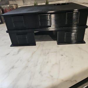 May include: A black, two-story miniature house model with rectangular windows. The model has a flat roof and is made of a solid, dark material. The windows are recessed, adding depth to the design. The house sits on a white marble surface.