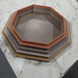 May include: A stack of four multi-colored, octagonal trays. The trays are in shades of gold, gray, and orange. The trays are nested inside each other, with the largest tray at the bottom and the smallest at the top. The trays are made of a solid material.
