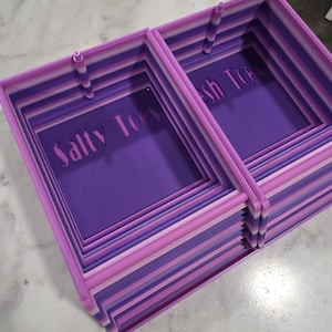 May include: Two rectangular, nested storage trays featuring a gradient of purple and pink horizontal stripes. The base of each tray is a solid dark purple with the text "Salty Toes" embossed in a lighter purple font on the interior floor.