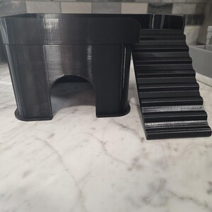 May include: A black 3D-printed animal hideout featuring a rectangular platform with a smooth top surface, a single arched entrance on the front side, and a set of integrated stairs leading up to the platform level from the right side.