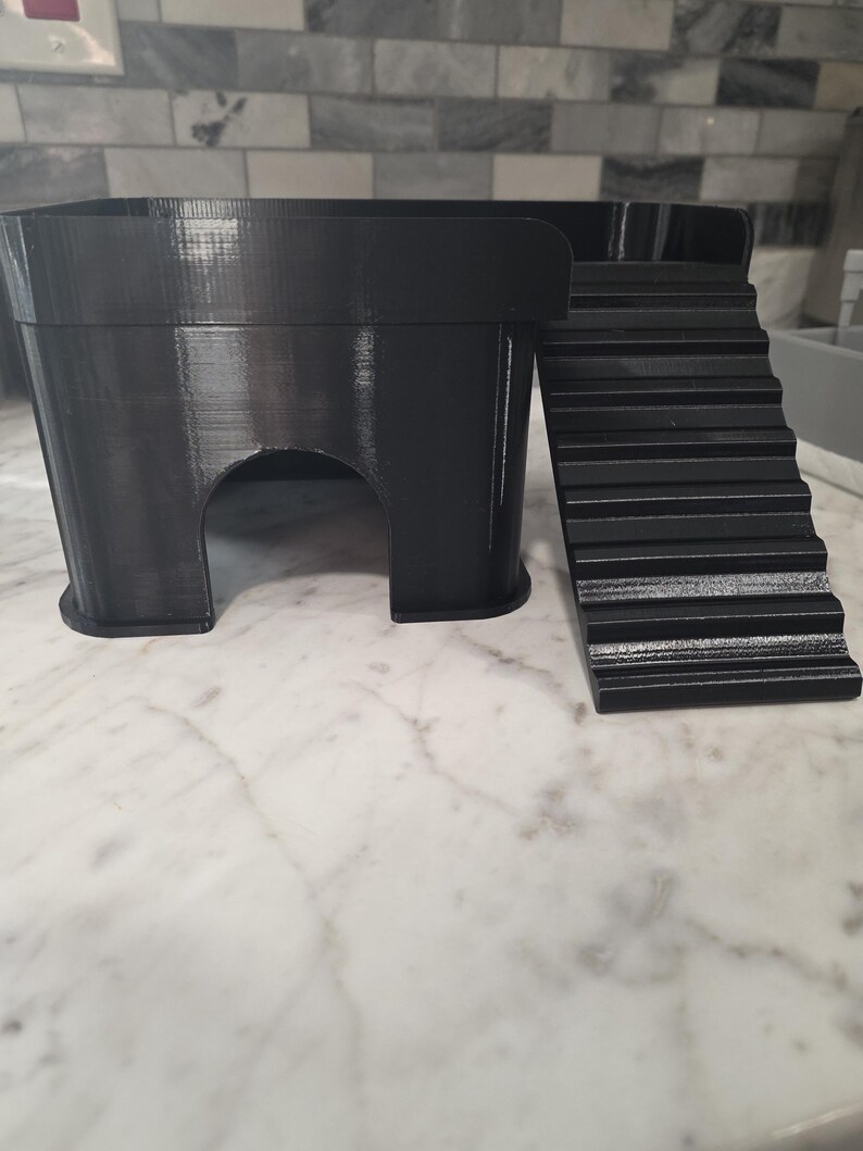 May include: A black 3D-printed animal hideout featuring a rectangular platform with a smooth top surface, a single arched entrance on the front side, and a set of integrated stairs leading up to the platform level from the right side.