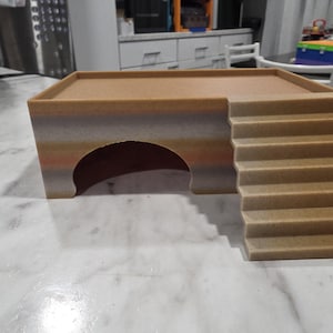 Puede incluir: Un refugio para mascotas de color tostado y degradado que presenta una plataforma superior plana, una entrada en arco semicircular en la parte frontal y un conjunto de escaleras integradas que conducen a la plataforma, con acabado mate texturizado.