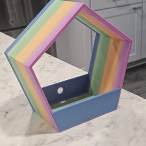 May include: A multi-colored, geometric storage box with a rainbow gradient on the sides and a solid blue base. The box has a pentagonal shape with an open top and a circular hole on one side.