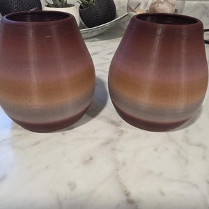 May include: Two brown and orange gradient-colored vases with a teardrop shape. The vases are sitting on a white marble surface. In the background, there are several small potted plants and a glass terrarium.