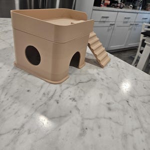 May include: A beige, two-story small animal hideout featuring a square base with a circular side opening and an arched front entrance. A set of stairs is attached to the side, leading to an open-top platform with a low railing on the upper level.