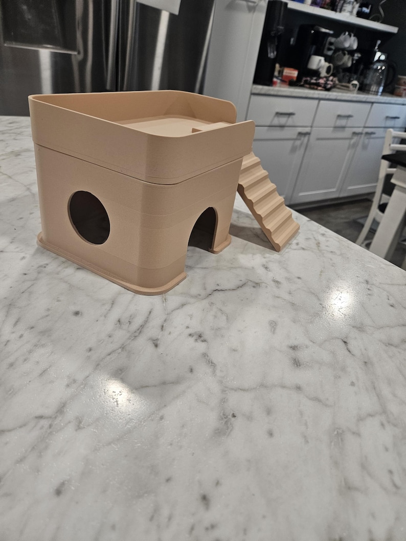 May include: A beige, two-story small animal hideout featuring a square base with a circular side opening and an arched front entrance. A set of stairs is attached to the side, leading to an open-top platform with a low railing on the upper level.