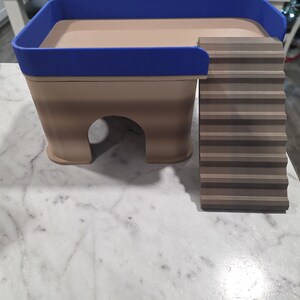 May include: A tan and brown pet hideout featuring a blue railing around the top platform and a set of stairs leading up to the platform. The structure has a hollow arched opening at the base for entry and exit, constructed with a layered design.