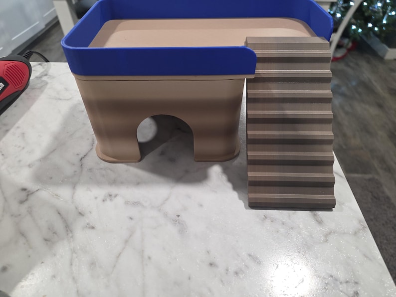 May include: A tan and brown pet hideout featuring a blue railing around the top platform and a set of stairs leading up to the platform. The structure has a hollow arched opening at the base for entry and exit, constructed with a layered design.