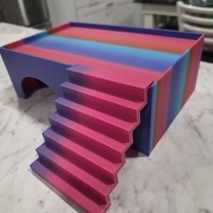 Puede incluir: Un escondite rectangular para mascotas con una plataforma superior plana y una escalera que conduce a ella. La estructura presenta un degradado vibrante de rayas rosas, moradas y azules. Una abertura arqueada es visible en el lateral.