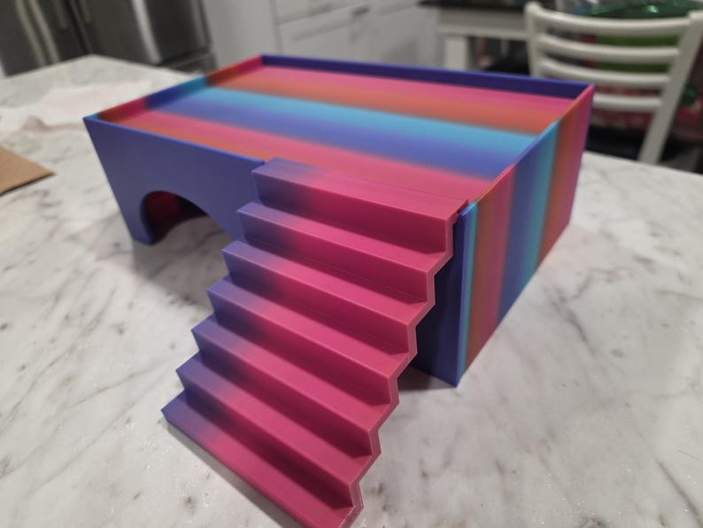 Puede incluir: Un escondite rectangular para mascotas con una plataforma superior plana y una escalera que conduce a ella. La estructura presenta un degradado vibrante de rayas rosas, moradas y azules. Una abertura arqueada es visible en el lateral.
