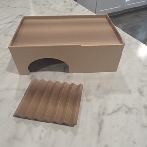 Puede incluir: Un recinto rectangular de color beige claro con una &uacute;nica abertura arqueada en la parte frontal y una pieza cuadrada separada a juego, de color beige claro, con una serie de crestas paralelas en zigzag en su superficie superior.