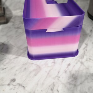 May include: A rectangular storage container featuring a gradient color pattern that transitions from deep purple to bright pink and light pink. The container has a lid with a raised edge and a small notch on the front side for opening the lid.