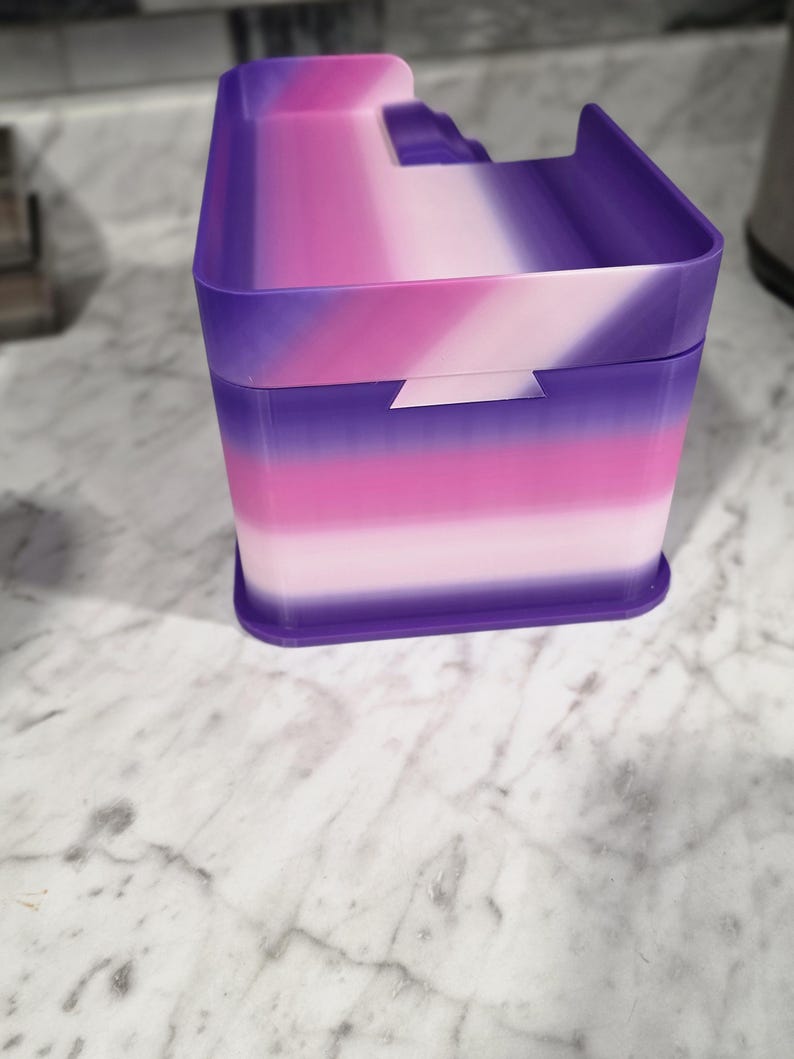 May include: A rectangular storage container featuring a gradient color pattern that transitions from deep purple to bright pink and light pink. The container has a lid with a raised edge and a small notch on the front side for opening the lid.
