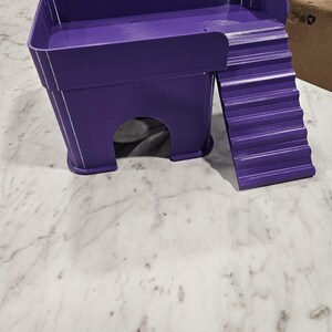 May include: A purple 3D-printed pet hideout featuring a raised platform with a surrounding wall and an attached staircase. The structure includes a hollow arched entrance at the base, providing a sheltered space for small animals to rest or climb.